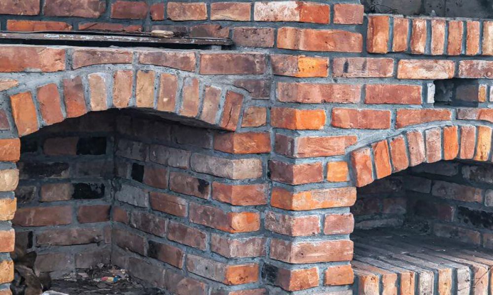 Brick oven structure with arched openings; located outdoors, the red and brown bricks form two separate oven chambers, set against a rustic environment with visible mortar joints.