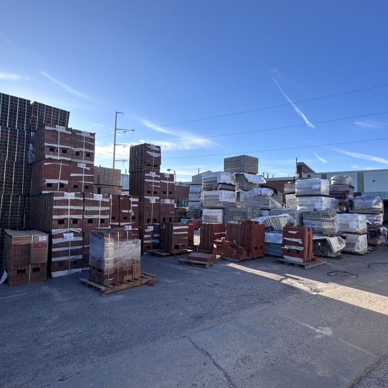A storage yard with stacks of bricks and building materials on wooden pallets. The sky is clear and blue, and there are power lines visible in the background. The ground is paved with asphalt.