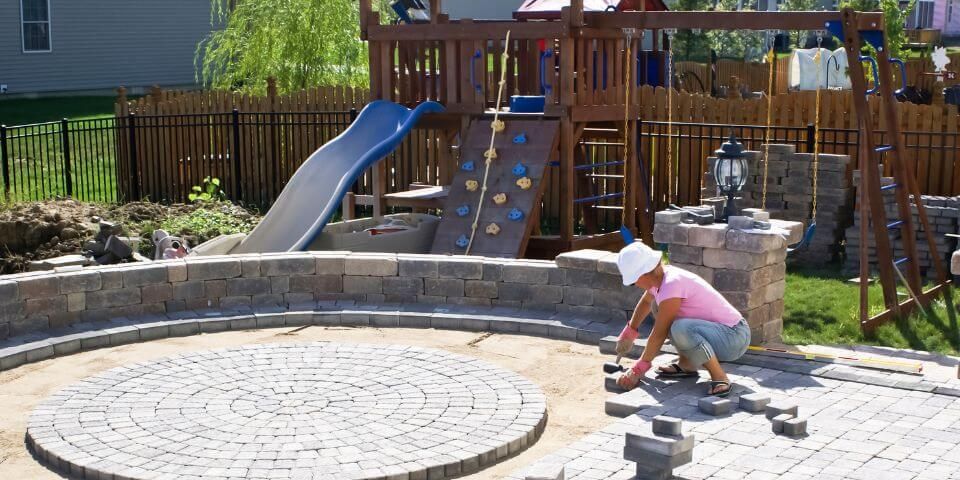 A person arranges bricks in a circular pattern on a patio, beside a playground with a slide, climbing wall, and swing set, surrounded by a fenced backyard.