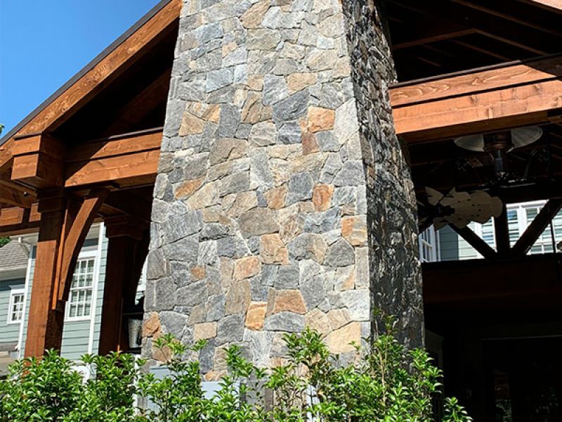 Outdoor patio with a large stone fireplace and wooden beams. Greenery surrounds the structure, and a ceiling fan is visible under the wooden roof. Text credits McHale Landscape Design.