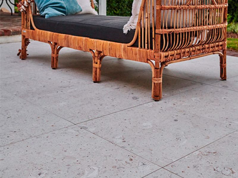A wicker bench with a black cushion and teal pillows is placed on a light gray tiled patio. The setting has a relaxed, outdoor ambiance.
