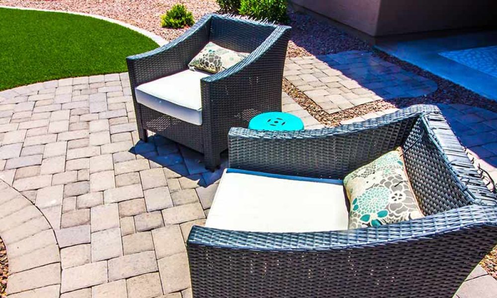Wicker patio chairs with cushions on a paved courtyard, adjacent to artificial turf, creating a cozy outdoor seating area.