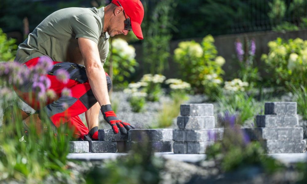 Man in red cap and gloves landscaping a garden with bricks, surrounded by green plants and flowers.