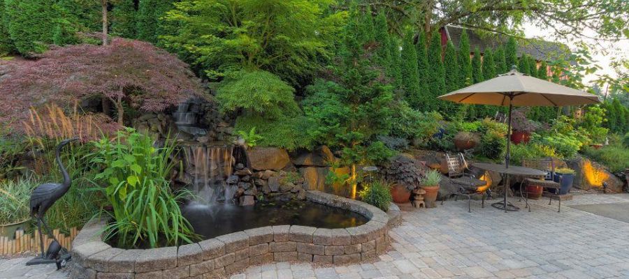 A landscaped garden with a waterfall, pond, and patio area featuring a fire pit, outdoor furniture, and an umbrella, surrounded by lush greenery and evening lighting.
