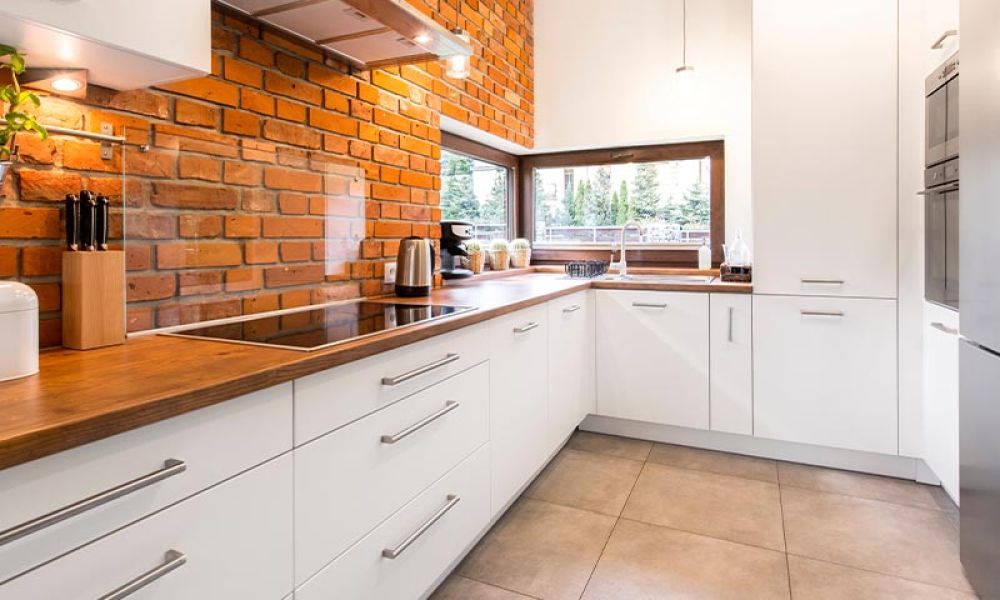 Modern kitchen with white cabinets, wood countertops, brick wall, large window, and stainless steel appliances.