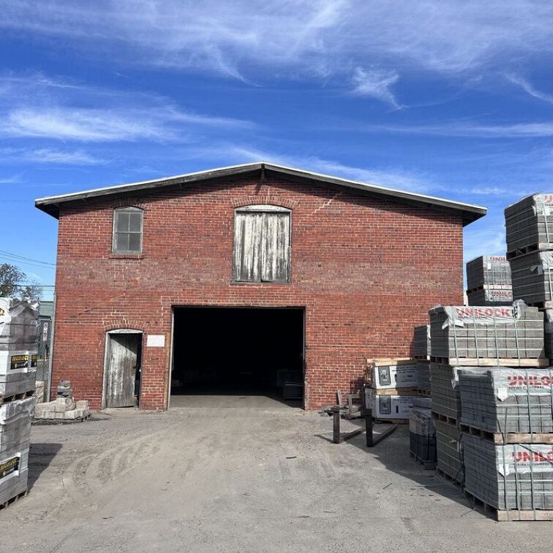 A red brick warehouse with a partially open door under a blue sky. Pallets of materials are stacked around the building on a dirt surface. There are some clouds in the sky.