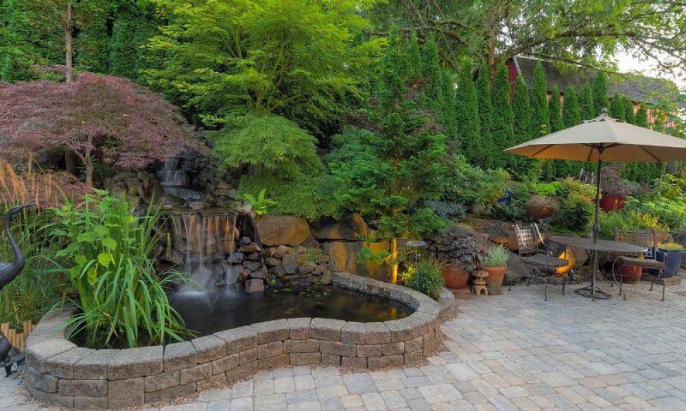 A landscaped garden with a waterfall, pond, and patio area featuring a fire pit, outdoor furniture, and an umbrella, surrounded by lush greenery and evening lighting.
