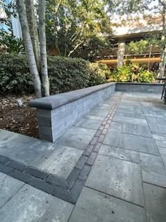 A stone pathway leads to a low retaining wall surrounded by trees and bushes. The area is shaded, and a building is visible in the background.