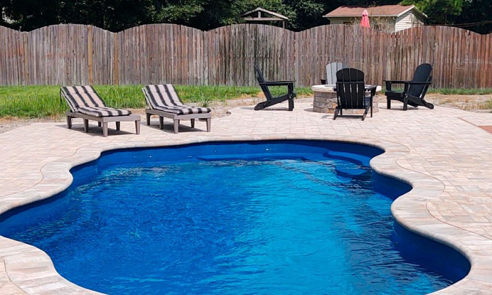 A backyard scene with a kidney-shaped swimming pool. Surrounding the pool are several lounge chairs with striped cushions and Adirondack chairs around a fire pit. A wooden fence encloses the area, and trees are visible in the background.