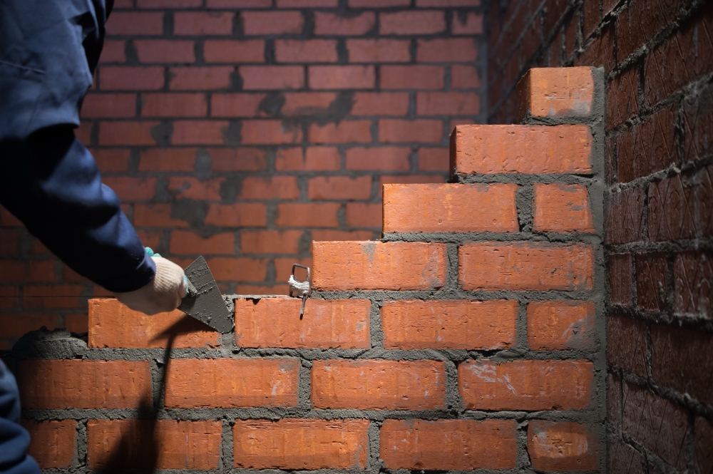 Builder laying bricks and applying mortar to create a sturdy wall in a dimly lit construction area.