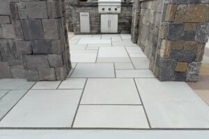 Modern stone patio with outdoor kitchen and grill, featuring sleek gray pavers and rustic stone walls.