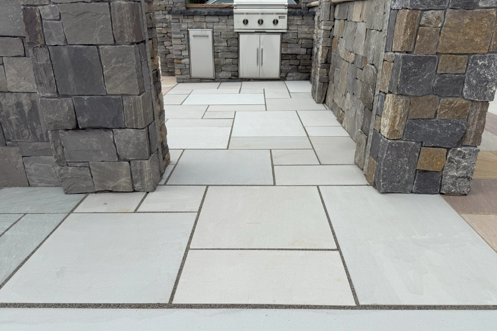 Modern stone patio with outdoor kitchen and grill, featuring sleek gray pavers and rustic stone walls.