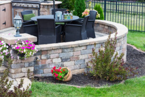 Outdoor patio with round table, wicker chairs, and brick walls, adorned by vibrant flowers and lush greenery.