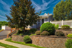 Charming suburban home with lush landscaping, brick retaining wall, and white picket fence under a clear blue sky.