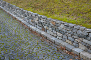 Stone wall on cobblestone path bordered by grassy hill, perfect for rustic landscape and garden design inspiration.