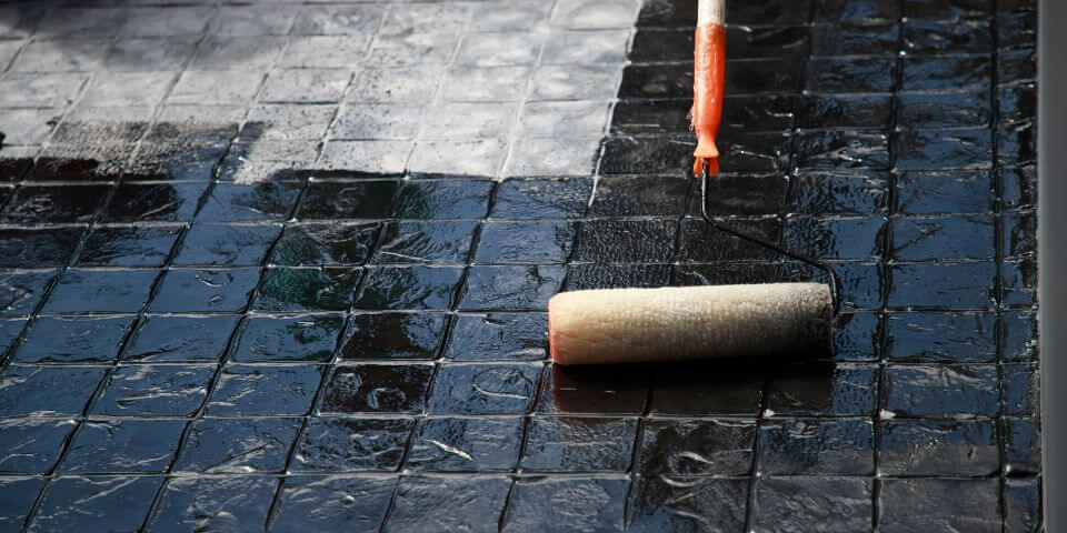 A paint roller applies a glossy black coating to textured tiles, reflecting a cloudy sky above.