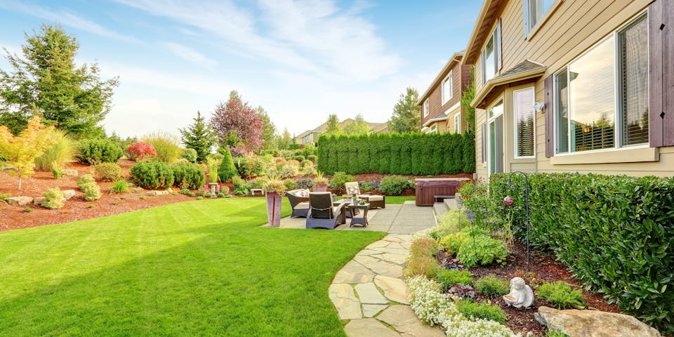A well-manicured backyard with a green lawn and a stone path leading to a patio with outdoor furniture, adjacent to a two-story house. Landscaping includes vibrant shrubs and trees.