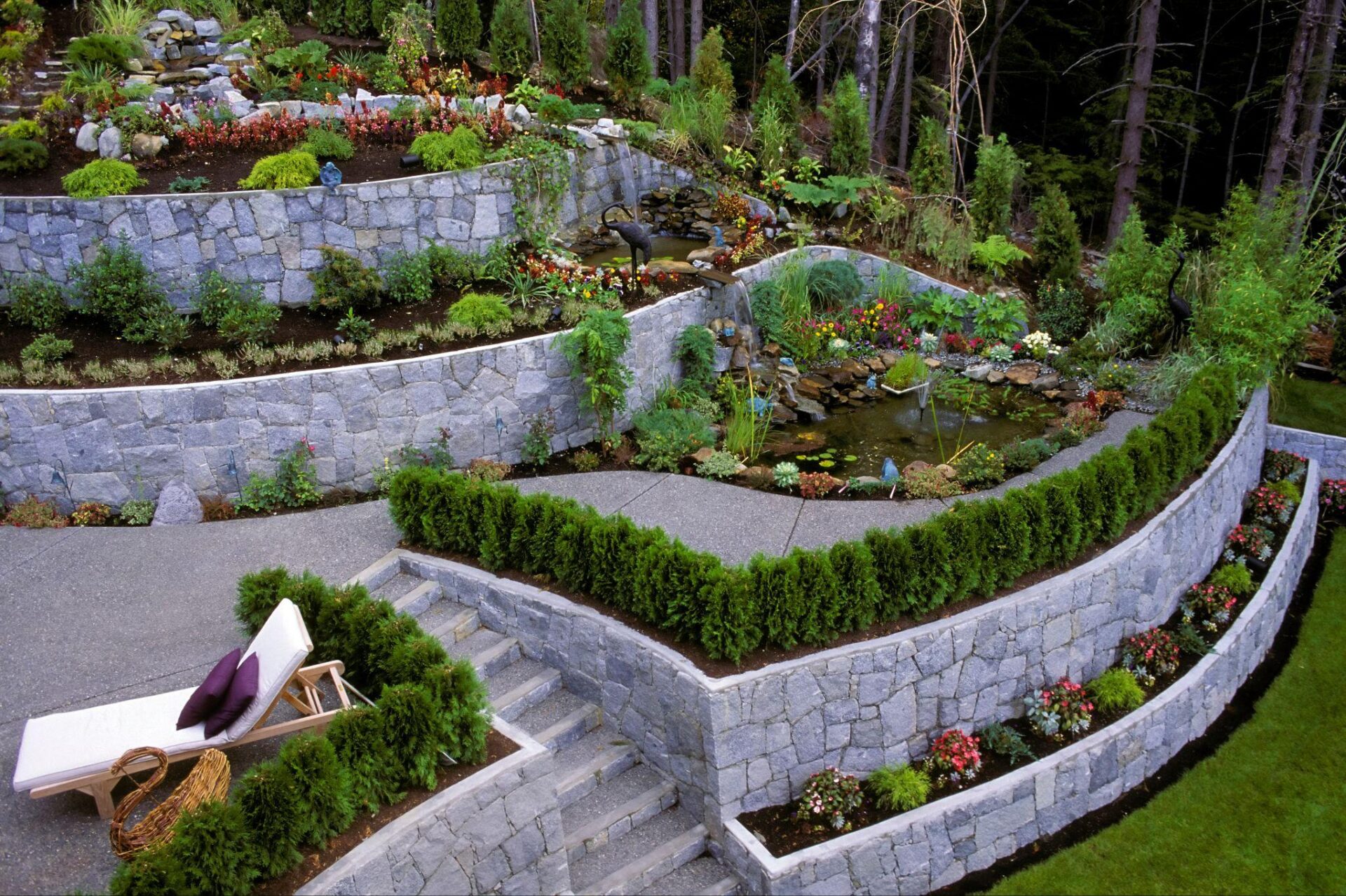 Landscaped garden with stone walls, lush greenery, colorful flowers, and cozy lounge chair. Relaxing outdoor retreat.