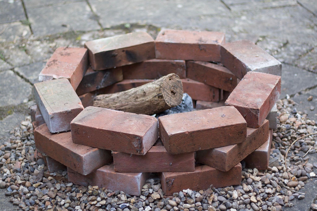DIY brick fire pit with wooden log in a pebble-filled outdoor setting.