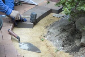 Worker laying bricks for garden pathway with trowel and rubber mallet, focusing on precision and alignment.