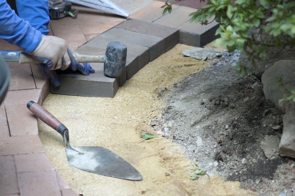 Worker laying bricks for garden pathway with trowel and rubber mallet, focusing on precision and alignment.