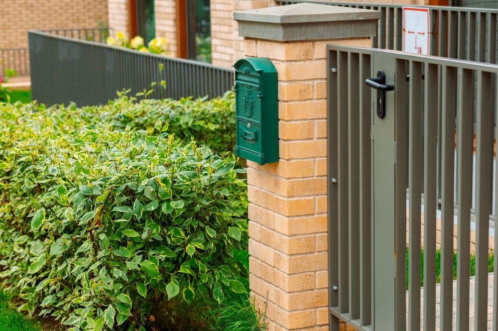Green mailbox on a brick pillar next to a metal gate and lush hedge in a residential setting.