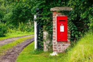 Red postbox on a brick pillar by a rural dirt path, surrounded by lush green foliage on a summer day.