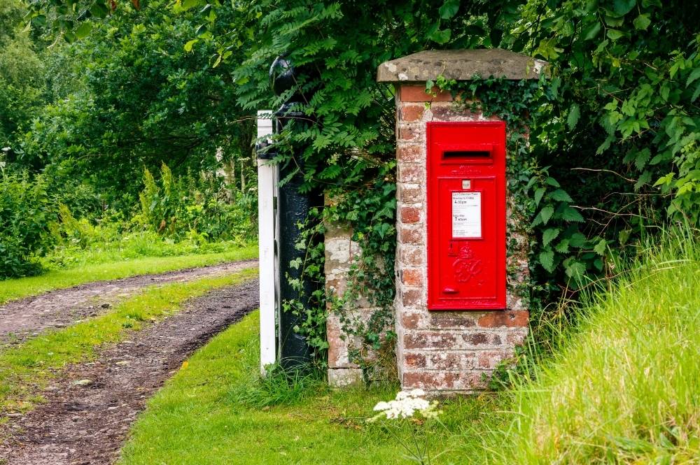 Red postbox on a brick pillar by a rural dirt path, surrounded by lush green foliage on a summer day.